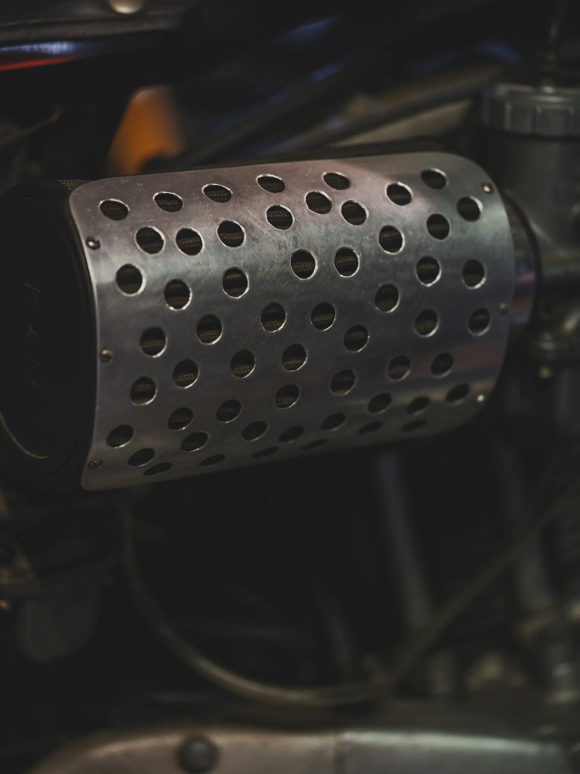 Macro detail of a hand-perforated titanium exhaust heat shield catching hard studio light